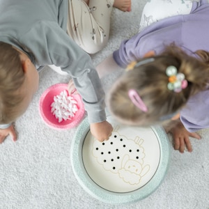 Könnte beinhalten: Zwei Kinder spielen ein Spiel mit einem weißen Keramikteller mit einem schwarz-weißen Schafdesign. Der Teller ist von einem hellblauen Rand umgeben. Die Kinder legen kleine weiße Pompons in den Teller.