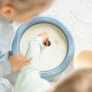 Könnte beinhalten: Zwei Kinder spielen mit einem blauen und weißen Holzspielzeug in Form eines Frosches. Der Frosch hat ein Loch im Maul und die Kinder benutzen einen Holzstab, um Gegenstände durch das Loch zu stecken.