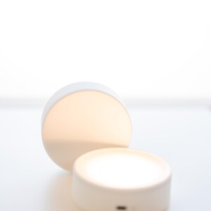 K&ouml;nnte beinhalten: Zwei wei&szlig;e, runde LED-Leuchten, sanft beleuchtet. Eine steht aufrecht, die andere ist geneigt, beide strahlen ein warmes Licht aus. Die Leuchten befinden sich auf einer wei&szlig;en Oberfl&auml;che, was auf ein minimalistisches Design hindeutet, m&ouml;glicherweise f&uuml;r die Inneneinrichtung oder Umgebungsbeleuchtung.