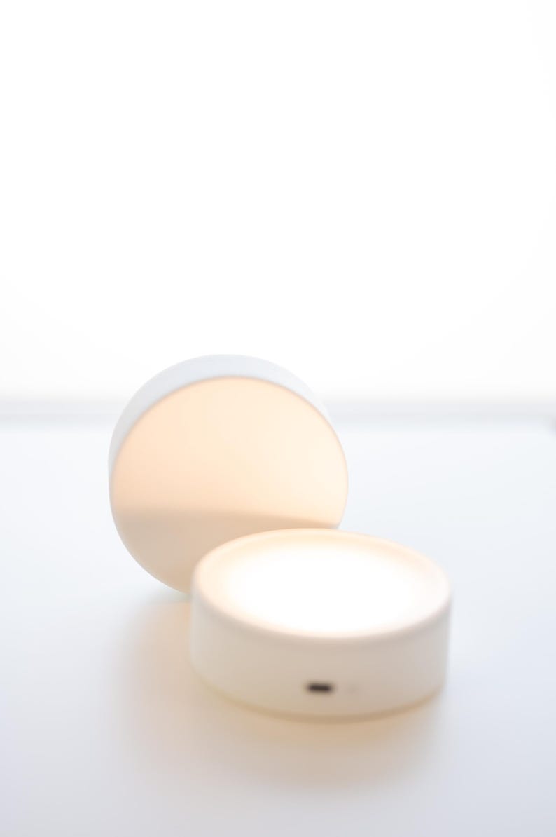 K&ouml;nnte beinhalten: Zwei wei&szlig;e, runde LED-Leuchten, sanft beleuchtet. Eine steht aufrecht, die andere ist geneigt, beide strahlen ein warmes Licht aus. Die Leuchten befinden sich auf einer wei&szlig;en Oberfl&auml;che, was auf ein minimalistisches Design hindeutet, m&ouml;glicherweise f&uuml;r die Inneneinrichtung oder Umgebungsbeleuchtung.