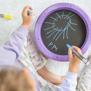 Könnte beinhalten: Ein Kind zeichnet mit einem blauen Stift auf einem runden, lila und schwarzen Schiefertafel. Die Schiefertafel hat einen lilafarbenen Rand und die Wörter "PA" sind darauf geschrieben.