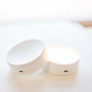 K&ouml;nnte beinhalten: Zwei wei&szlig;e, kreisf&ouml;rmige LED-Leuchten mit minimalistischem Design. Jede Leuchte hat eine flache Oberseite und einen zylindrischen Sockel. Eine Leuchte ist geneigt und zeigt einen kleinen, schwarzen Ladeanschluss. Die andere Leuchte leuchtet und strahlt ein sanftes Leuchten aus.