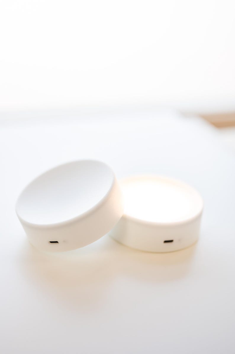 K&ouml;nnte beinhalten: Zwei wei&szlig;e, kreisf&ouml;rmige LED-Leuchten mit minimalistischem Design. Jede Leuchte hat eine flache Oberseite und einen zylindrischen Sockel. Eine Leuchte ist geneigt und zeigt einen kleinen, schwarzen Ladeanschluss. Die andere Leuchte leuchtet und strahlt ein sanftes Leuchten aus.