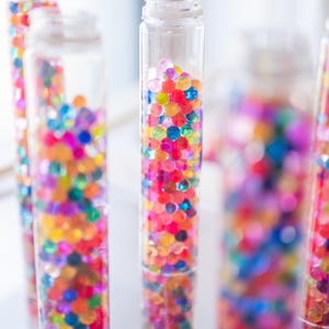 Könnte beinhalten: Vier durchsichtige Glasflaschen gefüllt mit bunten, durchsichtigen, runden Wasserperlen. Die Flaschen sind in einer Reihe auf einer weißen Oberfläche angeordnet.