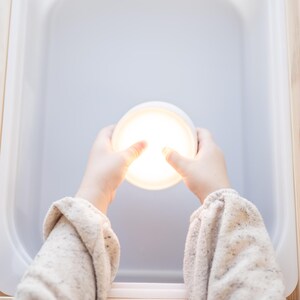 K&ouml;nnte beinhalten: Ein helles, rundes, wei&szlig;es Licht wird von zwei H&auml;nden gehalten, die den Raum beleuchten. Die H&auml;nde tragen einen beigen Pullover. Das Licht befindet sich in einem wei&szlig;en, rechteckigen Beh&auml;lter, der in einem Holzrahmen steht.