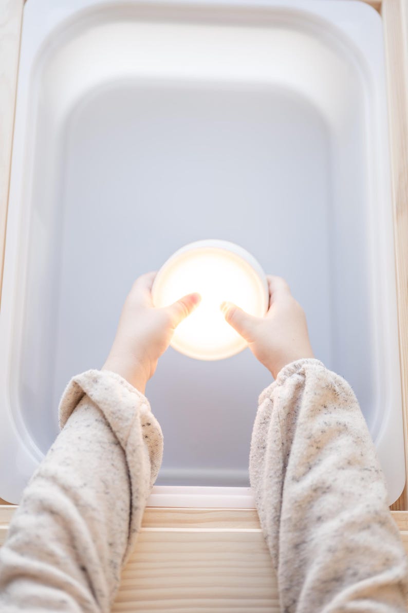 K&ouml;nnte beinhalten: Ein helles, rundes, wei&szlig;es Licht wird von zwei H&auml;nden gehalten, die den Raum beleuchten. Die H&auml;nde tragen einen beigen Pullover. Das Licht befindet sich in einem wei&szlig;en, rechteckigen Beh&auml;lter, der in einem Holzrahmen steht.