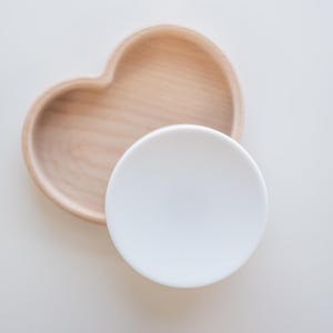K&ouml;nnte beinhalten: Eine herzf&ouml;rmige Holzschale und ein kleiner, runder wei&szlig;er Keramikteller. Die Holzschale hat eine nat&uuml;rliche Holzmaserung. Der wei&szlig;e Teller ist glatt und liegt vor der Holzschale.