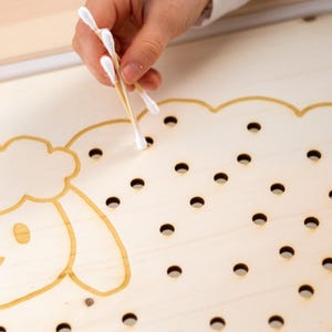 Könnte beinhalten: Ein Holzbrett mit einer Schafform und Löchern für eine sensorische Aktivität. Eine Hand verwendet einen Wattestäbchen, um die Löcher zu füllen.