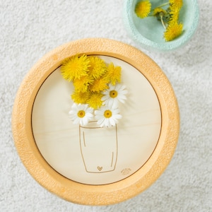 Könnte beinhalten: Ein Holz-Untersetzer mit einem hellbraunen Außenring und einer weißen Mitte. Die Mitte zeigt eine Zeichnung einer Vase mit einem Herzen und einem Strauß gelber und weißer Blumen.