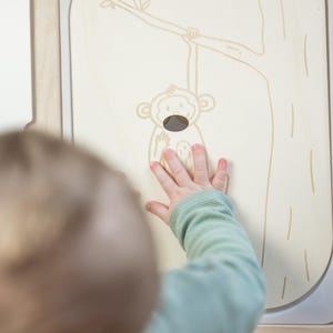 Könnte beinhalten: Ein Kind streckt die Hand aus, um ein Holzpaneel mit einer Zeichnung eines Affen zu berühren, der an einem Ast hängt. Das Paneel ist Teil einer Holzspielstruktur.