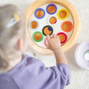 Könnte beinhalten: Ein Holzspielzeug mit einer runden Basis und einer weißen Mitte. Die Basis hat 10 Löcher am Rand, jedes mit einem farbigen Ring. Eine Kinderhand greift in eines der Löcher.