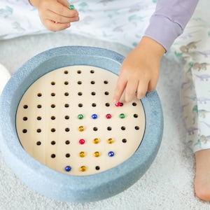 Könnte beinhalten: Eine Kinderhand legt eine rote Murmel in ein Holz-Steckspiel mit blauem Rand. Das Spielfeld hat verschiedene farbige Murmeln in den Löchern.