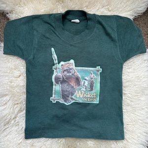 May include: Dark green t-shirt featuring a graphic of an Ewok character holding a spear, with the text "Wicket the Ewok" in a frame. The shirt is made of a soft material and has a crew neck.
