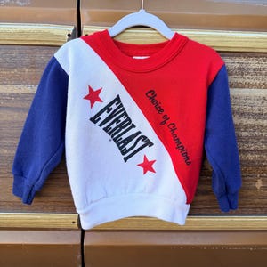 May include: A vintage Everlast sweatshirt in red, white, and blue. The long-sleeved top features the Everlast logo and the phrase "Choice of Champions." The sleeves are blue, and the body is divided diagonally with red and white sections.