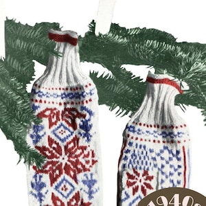 May include: Two white knitted mitten ornaments with red and blue snowflake patterns, hanging from a green pine branch with two white candles.