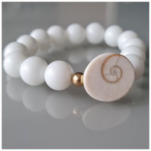 Peut inclure: Un bracelet de perles blanches avec un grand charme en forme de coquillage blanc cassé avec un motif en spirale doré. Le bracelet comprend une perle dorée.