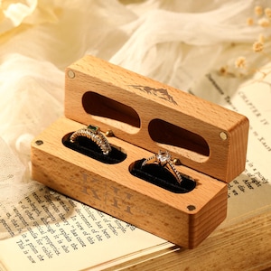 Könnte beinhalten: Eine Holzringbox mit zwei Fächern, die jeweils einen Ring enthalten. Ein Ring hat einen grünen Stein, der andere einen klaren Stein. Die Box ist geöffnet und zeigt die Ringe auf einem schwarzen Samtfutter. Die Box liegt auf einem offenen Buch.