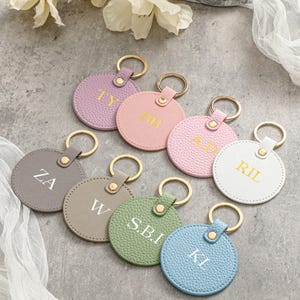 Könnte beinhalten: Eine Sammlung personalisierter Schlüsselanhänger in verschiedenen Pastellfarben, darunter Lavendel, Rosa und Blau. Jeder runde Schlüsselanhänger hat einen goldfarbenen Ring und ist mit Initialen in einer Kontrastfarbe geprägt. Die Schlüsselanhänger sind auf einer grauen Oberfläche mit weißem Stoff angeordnet.