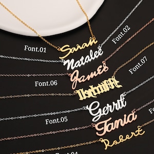 Könnte beinhalten: Sieben goldene Halsketten mit verschiedenen Schriftarten und Namen, darunter Sarah, Natalie, James, Isidora, Gerrit, Tania und Robert. Die Halsketten werden auf einer weißen Oberfläche mit schwarzem Hintergrund präsentiert.