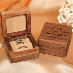 Puede incluir: Dos cajas de anillos de madera. Una caja está abierta y tiene dos anillos dentro. La otra caja está cerrada y tiene el texto "Sarah & Robert 18.06.2023" grabado en la parte superior.