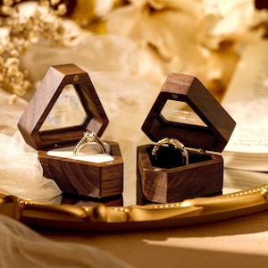 May include: Two wooden ring boxes with clear glass lids. One box has a gold ring with diamonds inside, and the other box has a gold band ring inside.