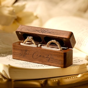 Könnte beinhalten: Eine Holzkiste für Ringe mit einem Scharnierdeckel, graviert mit den Worten "The Candy". Die Kiste ist geöffnet und zeigt zwei goldene Ringe mit Diamanten. Die Kiste steht auf einem Buch.