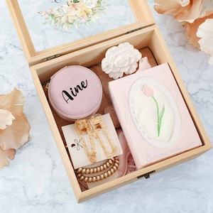Könnte beinhalten: Eine Holzkiste gefüllt mit Geschenken, darunter eine runde rosa Box mit dem Wort "Aime" darauf, goldener Schmuck, eine rosa und weiße Blume und ein weißes Taschentuch mit einem rosa Blumenmuster.