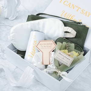 May include: A white gift box containing a green fabric pouch, a white sleep mask, a white tumbler with the name "Usenia" on it, a card that says "Will you help me tie the knot?" and a small bouquet of yellow flowers in a green pouch. The box lid has the text "I can't say I do without you" in gold lettering.