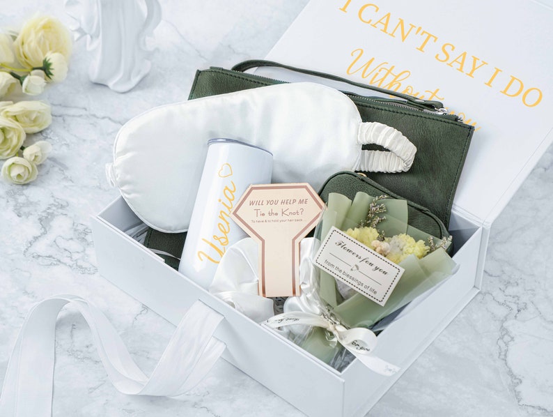 May include: A white gift box containing a green fabric pouch, a white sleep mask, a white tumbler with the name "Usenia" on it, a card that says "Will you help me tie the knot?" and a small bouquet of yellow flowers in a green pouch. The box lid has the text "I can't say I do without you" in gold lettering.