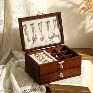 May include: A wooden jewellery box with a hinged lid and two drawers. The lid is open and reveals compartments for storing earrings, rings, and other jewellery. The box is brown and has a heart-shaped design on the front.