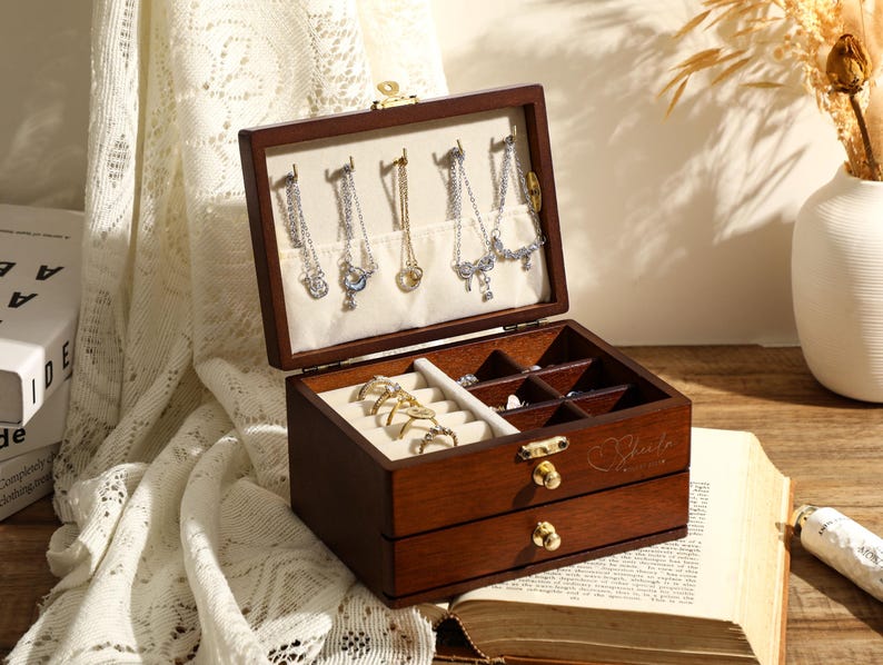 May include: A wooden jewellery box with a hinged lid and two drawers. The lid is open and reveals compartments for storing earrings, rings, and other jewellery. The box is brown and has a heart-shaped design on the front.