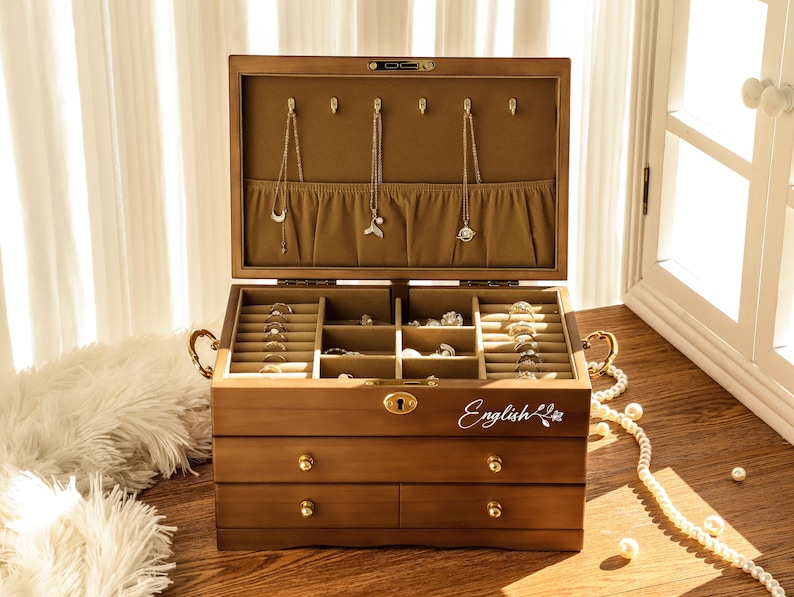 May include: A wooden jewelry box with multiple compartments and drawers. The box is open, revealing necklaces and rings. The box has a light brown finish, gold hardware, and the word "English" is written on the front.