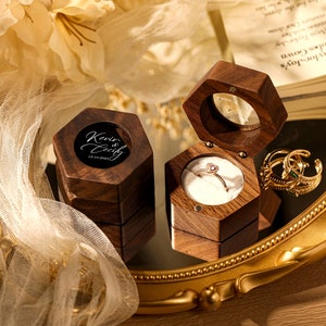 Puede incluir: Una caja de anillo de madera hexagonal con una tapa transparente. La tapa tiene una inscripción negra que dice "Kevin & Cathy 12.12.2022". La caja está abierta y hay un anillo dentro.