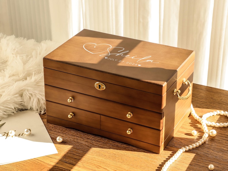 May include: A wooden jewelry box with three drawers and a hinged lid. The box is a warm brown color with gold-colored hardware. The lid is engraved with the name "Sheila", a heart, and the date "July 27, 2026".