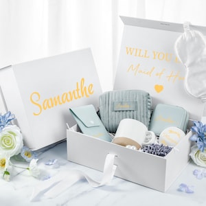 May include: A white gift box with a blue and white patterned interior. The box contains a sleep mask, a small blue pouch, a white mug, a small blue pouch, and a small white box. The box is labeled "Will you be my Maid of Honor?" and "Samantha".