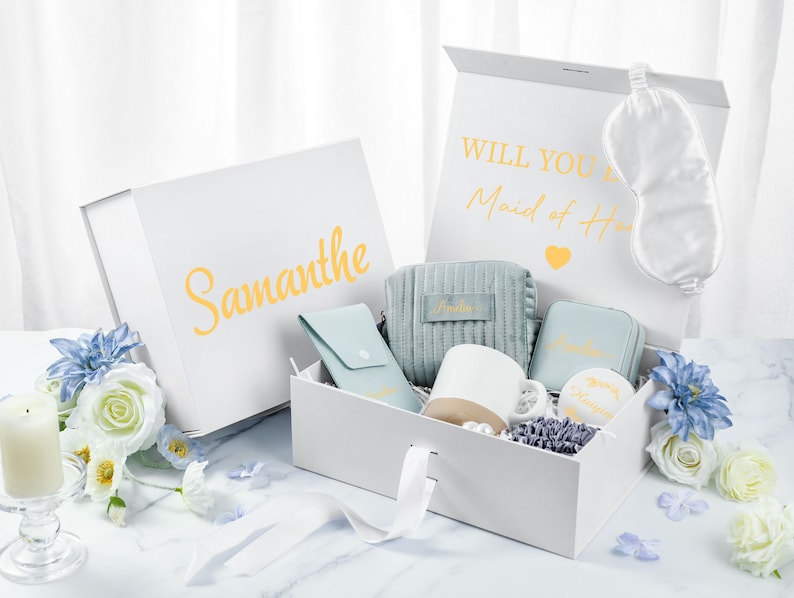 May include: A white gift box with a blue and white patterned interior. The box contains a sleep mask, a small blue pouch, a white mug, a small blue pouch, and a small white box. The box is labeled "Will you be my Maid of Honor?" and "Samantha".