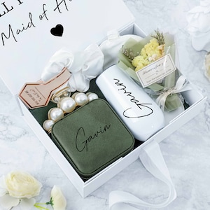 May include: A white gift box with a white ribbon tied around it. The box contains a green pouch with the name "Gavin" written on it, a white tumbler with the name "Gavin" written on it, a small bouquet of dried flowers, and a hair scrunchie. The box has the text "Will you be my Maid of Honor?" written on the top.