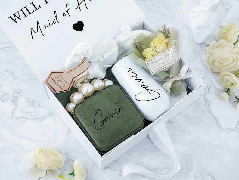 May include: A white gift box with a white ribbon tied around it. The box contains a green pouch with the name "Gavin" written on it, a white tumbler with the name "Gavin" written on it, a small bouquet of dried flowers, and a hair scrunchie. The box has the text "Will you be my Maid of Honor?" written on the top.