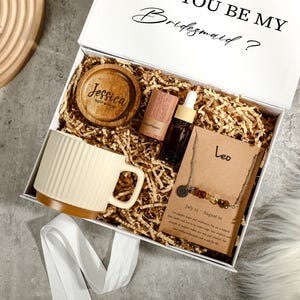 Può includere: Una scatola regalo bianca con la scritta "YOU BE MY Bridesmaid?" contiene una tazza, un sottobicchiere, una bottiglia di olio essenziale e una collana. La tazza è color crema con base marrone. Il sottobicchiere è in legno con il nome "Jessica". La collana ha un ciondolo Leone.