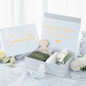 May include: White gift box with a gold inscription that reads "Will you be my Maid of Honor." The box is open and contains a smaller green box with the name "Ashley" on it, a white mug with the name "Ashley" on it, a small bottle of lotion, and a small bouquet of white flowers. The box is sitting on a marble surface.