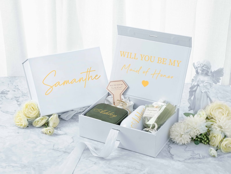 May include: White gift box with a gold inscription that reads "Will you be my Maid of Honor." The box is open and contains a smaller green box with the name "Ashley" on it, a white mug with the name "Ashley" on it, a small bottle of lotion, and a small bouquet of white flowers. The box is sitting on a marble surface.