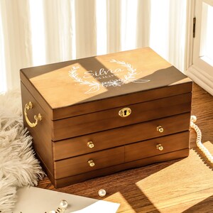 May include: A wooden jewelry box with three drawers and a hinged lid. The box is a warm brown color with gold-colored hardware. The lid is engraved with the name "Silvia" and a floral design. The box is sitting on a wooden surface.