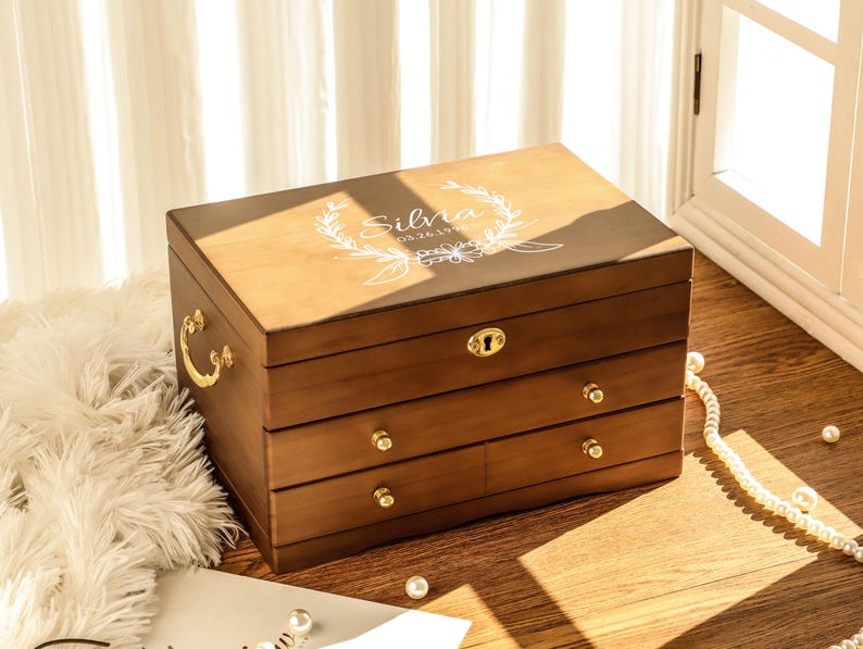 May include: A wooden jewelry box with three drawers and a hinged lid. The box is a warm brown color with gold-colored hardware. The lid is engraved with the name "Silvia" and a floral design. The box is sitting on a wooden surface.