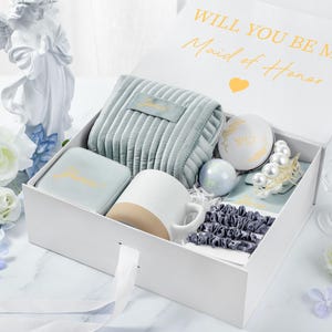 May include: A white gift box with a blue and white patterned interior. The box contains a variety of gifts, including a mug, a small pouch, a jewelry box, and a small decorative item. The box is open and the lid is propped up, revealing the contents. The lid of the box has the text "Will you be my Maid of Honor" in gold lettering.