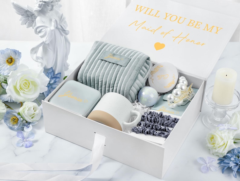 May include: A white gift box with a blue and white patterned interior. The box contains a variety of gifts, including a mug, a small pouch, a jewelry box, and a small decorative item. The box is open and the lid is propped up, revealing the contents. The lid of the box has the text "Will you be my Maid of Honor" in gold lettering.