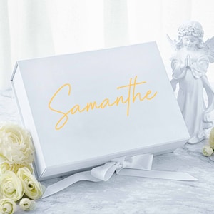 May include: White gift box with a satin ribbon and the name "Samantha" written in gold lettering. The box is sitting on a marble surface with white flowers.