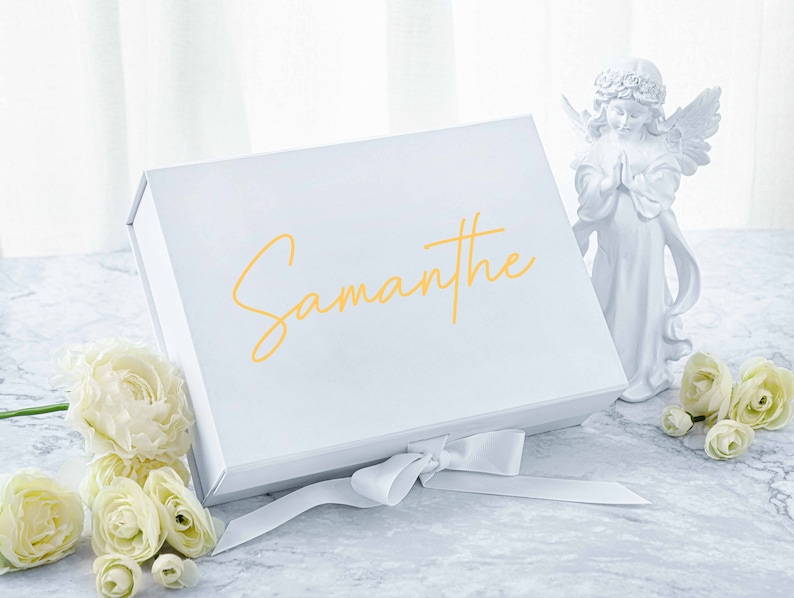 May include: White gift box with a satin ribbon and the name "Samantha" written in gold lettering. The box is sitting on a marble surface with white flowers.