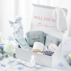 May include: A white gift box with a blue and white patterned interior. The box contains a white mug, a blue eye mask, a blue cosmetic bag, a blue jewelry box, and a round white trinket box. The box is decorated with the text "Will you be Bridesmaid?" and the words "Callie" and "Shirley" are written on the items inside the box.