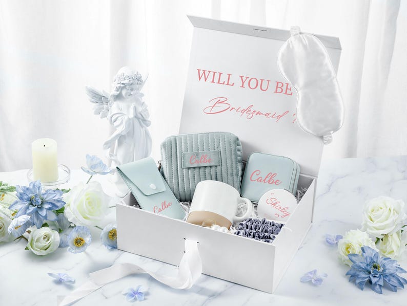 May include: A white gift box with a blue and white patterned interior. The box contains a white mug, a blue eye mask, a blue cosmetic bag, a blue jewelry box, and a round white trinket box. The box is decorated with the text "Will you be Bridesmaid?" and the words "Callie" and "Shirley" are written on the items inside the box.