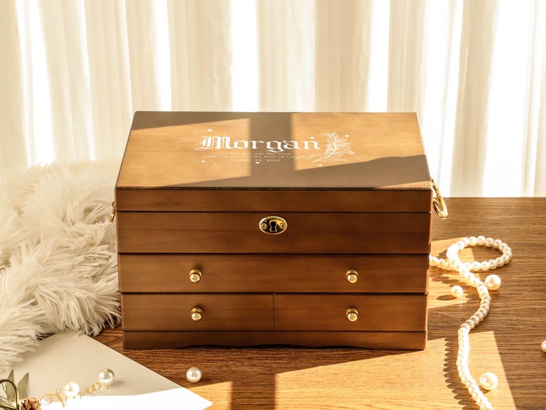 May include: A wooden jewelry box with three drawers and a hinged top. The box is a warm brown color with gold-colored hardware and the name "Morgan" in a decorative font on the lid. A pearl necklace and other decorative items are on the table.
